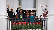 Barack and Michelle Obama, their children, and her mother, along with a costumed Easter Bunny, on a balcony waving.