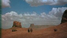 A color screenshot of several men on horses, in three groups: Native Americans on the left, Americans in the center, and Native Americans on the right. The men are located in a desert and in the background are large rock formations and blue skies with white clouds.