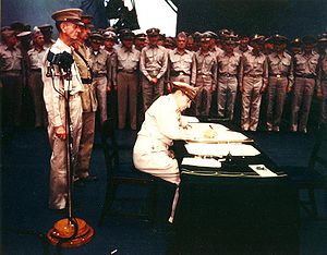 MacArthur is seated a small desk, writing. Two men in uniform stand behind him. A large crowd of men in uniform look on.