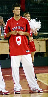 A man, wearing white pants and a red t-shirt with the word «RAPTORS» on the front, is standing on a basketball court.